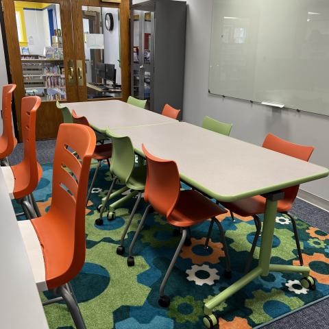 Image of Makerspace. The room has two movable tables, four table height chairs, a large whiteboard, and four counter height stools set before a fixed height counter. The room has swinging glass doors that face the Children's area.