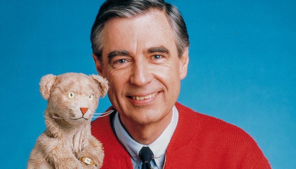 Photo of Mr. Rogers in his red cardigan and holding teddy bear puppet